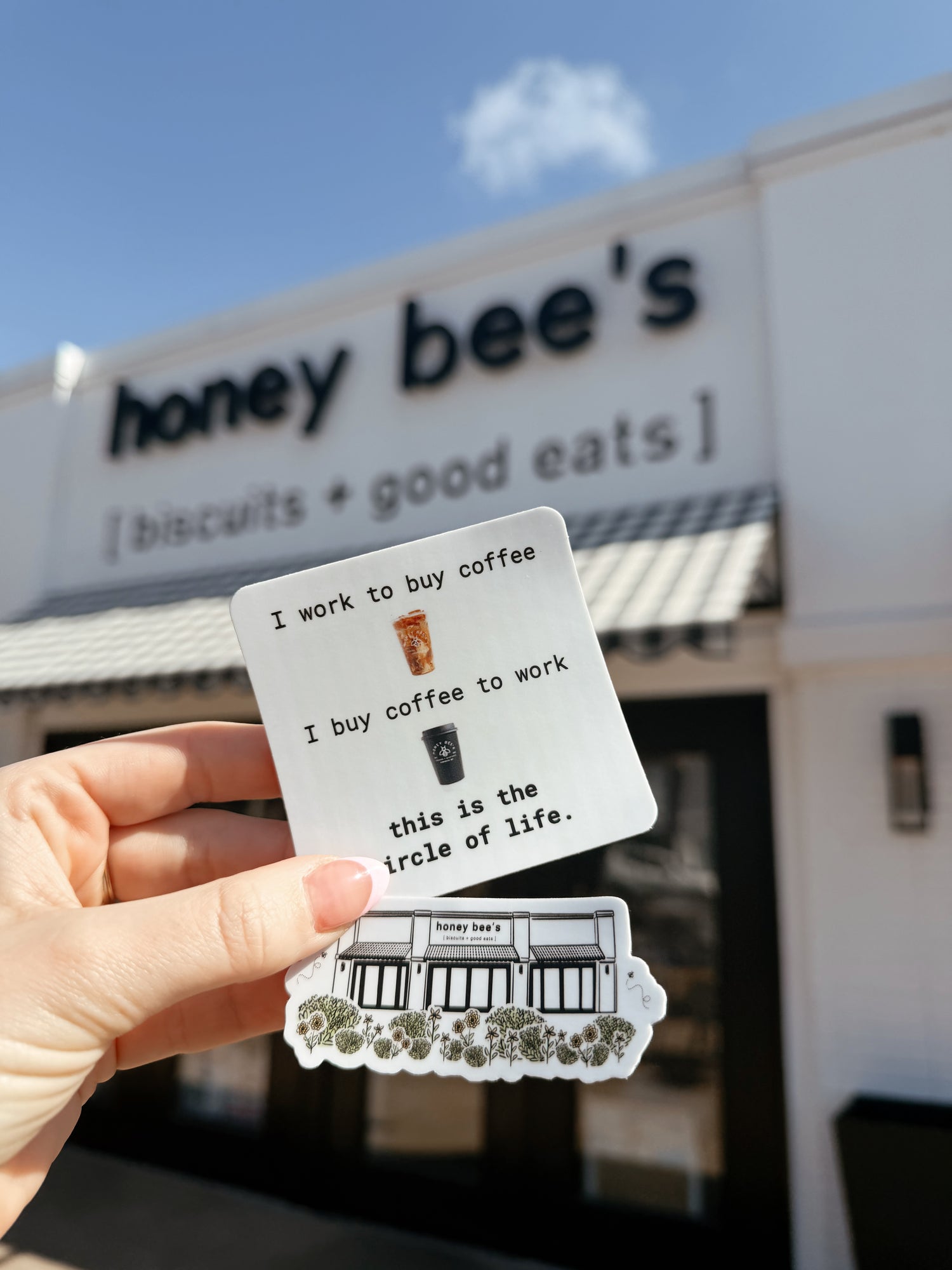 Person holding a card with humorous text about coffee is life in front of 'honey bee's' building